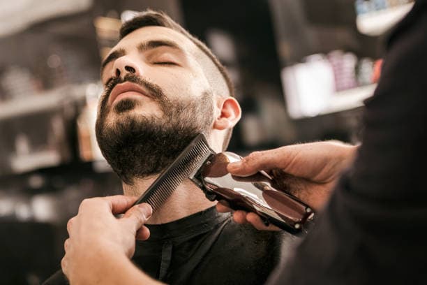 Barber trims a man's beard with electric clippers and a comb in a barbershop.